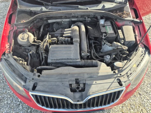 A top-down view of the engine bay in a red ŠKODA OCTAVIA 1.2 BENZIN/TNG, displaying various engine components, hoses, and the battery, with visible dust and grime on most surfaces.