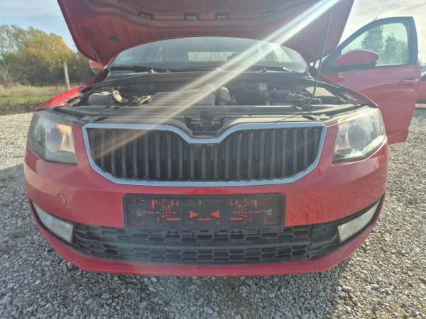 A ŠKODA OCTAVIA 1.2 BENZIN/TNG in red with its hood and front driver-side door open is parked on gravel under a sunny sky, displaying its engine.