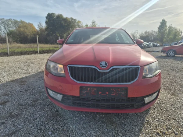 A ŠKODA OCTAVIA 1.2 BENZIN/TNG in red is parked on gravel outdoors, facing the camera. There are trees and several other cars in the background under a partly cloudy sky, with sunlight causing glare across the photo.