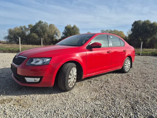 A ŠKODA OCTAVIA 1.2 BENZIN/TNG sedan, painted red, is parked outdoors on gravel with trees and a blue sky behind it, angled to show the front and driver’s side.