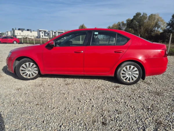 A ŠKODA OCTAVIA 1.2 BENZIN/TNG sedan is parked on gravel, with trees and modern buildings in the background under a clear blue sky.