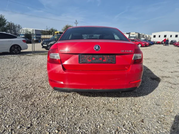 A ŠKODA OCTAVIA 1.2 BENZIN/TNG in red is parked on a gravel lot, photographed from behind with an empty license plate area. Other cars and buildings appear in the background beneath a blue sky.
