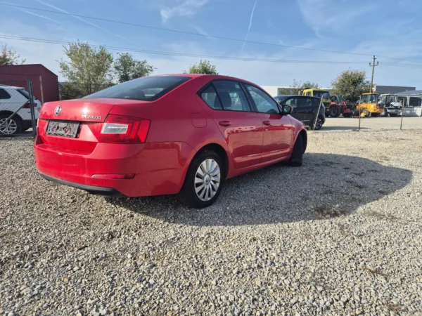 A ŠKODA OCTAVIA 1.2 BENZIN/TNG in red is parked on a gravel lot under a clear sky, with trees, fencing, and construction vehicles visible in the background.