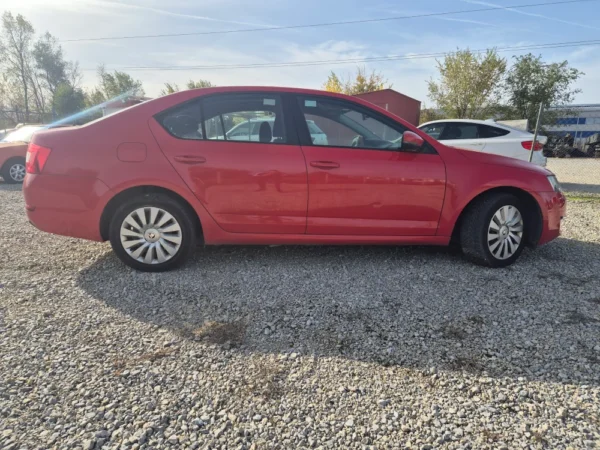 A ŠKODA OCTAVIA 1.2 BENZIN/TNG is parked on a gravel lot with trees, a white car, and a red building in the background on a sunny day.