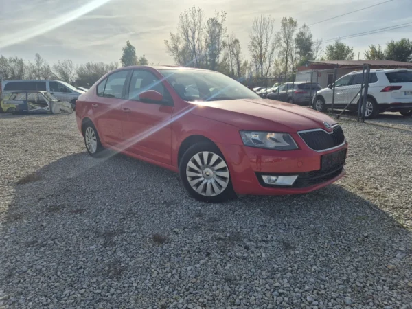 A ŠKODA OCTAVIA 1.2 BENZIN/TNG sedan in red is parked on a gravel lot under a sunny sky, with trees and other vehicles in the background. Sunlight causes a bright glare across the scene.