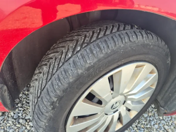 A close-up of the ŠKODA OCTAVIA 1.2 BENZIN/TNG's front wheel and tire, highlighting a Riken tire with a clear tread pattern. The red car is parked on gravel.