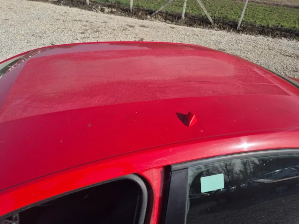A close-up of a ŠKODA OCTAVIA 1.2 BENZIN/TNG's dusty red roof with a small antenna near the center, part of an open door in the foreground, and gravel with fencing visible in the background.