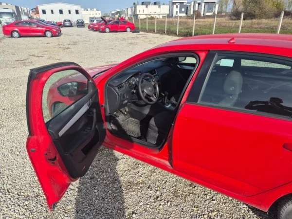 A ŠKODA OCTAVIA 1.2 BENZIN/TNG in red, parked on gravel with the driver-side door open; the steering wheel and front seats are visible, while other red cars and buildings appear in the background under a blue sky.