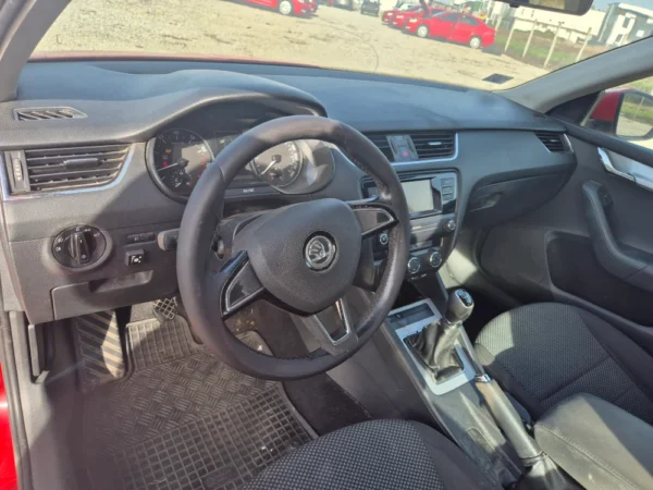Interior view of the ŠKODA OCTAVIA 1.2 BENZIN/TNG, featuring steering wheel, dashboard with gauges, gear stick, infotainment screen, air vents, and black upholstered front seats illuminated by sunlight through the windows.