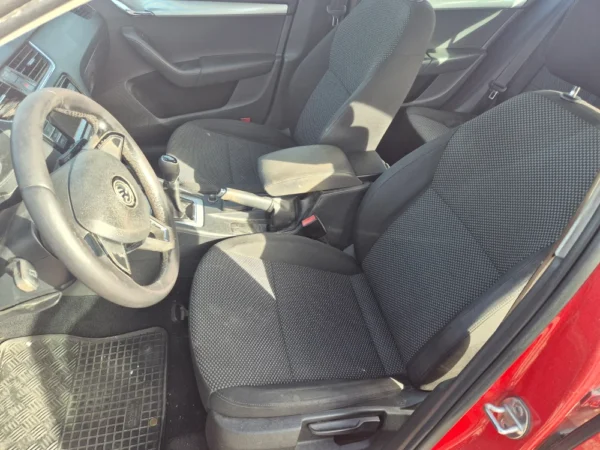 Photo of the front interior of a ŠKODA OCTAVIA 1.2 BENZIN/TNG, featuring two black fabric seats, steering wheel, center console with gear shift, armrest, and part of a red car door. Sunlight illuminates the seats.