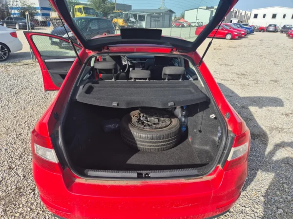 A red ŠKODA OCTAVIA 1.2 BENZIN/TNG is parked on gravel, trunk open to show a spare tire and tools inside, with the rear left door also open, surrounded by other cars and industrial buildings in the background.